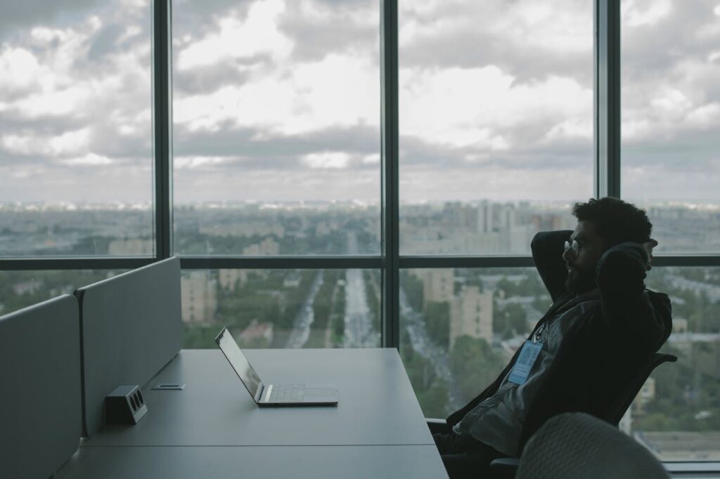 pexels photo 5483064 A man sits in an office with hands on head in front of a laptop, overlooking a cityscape.